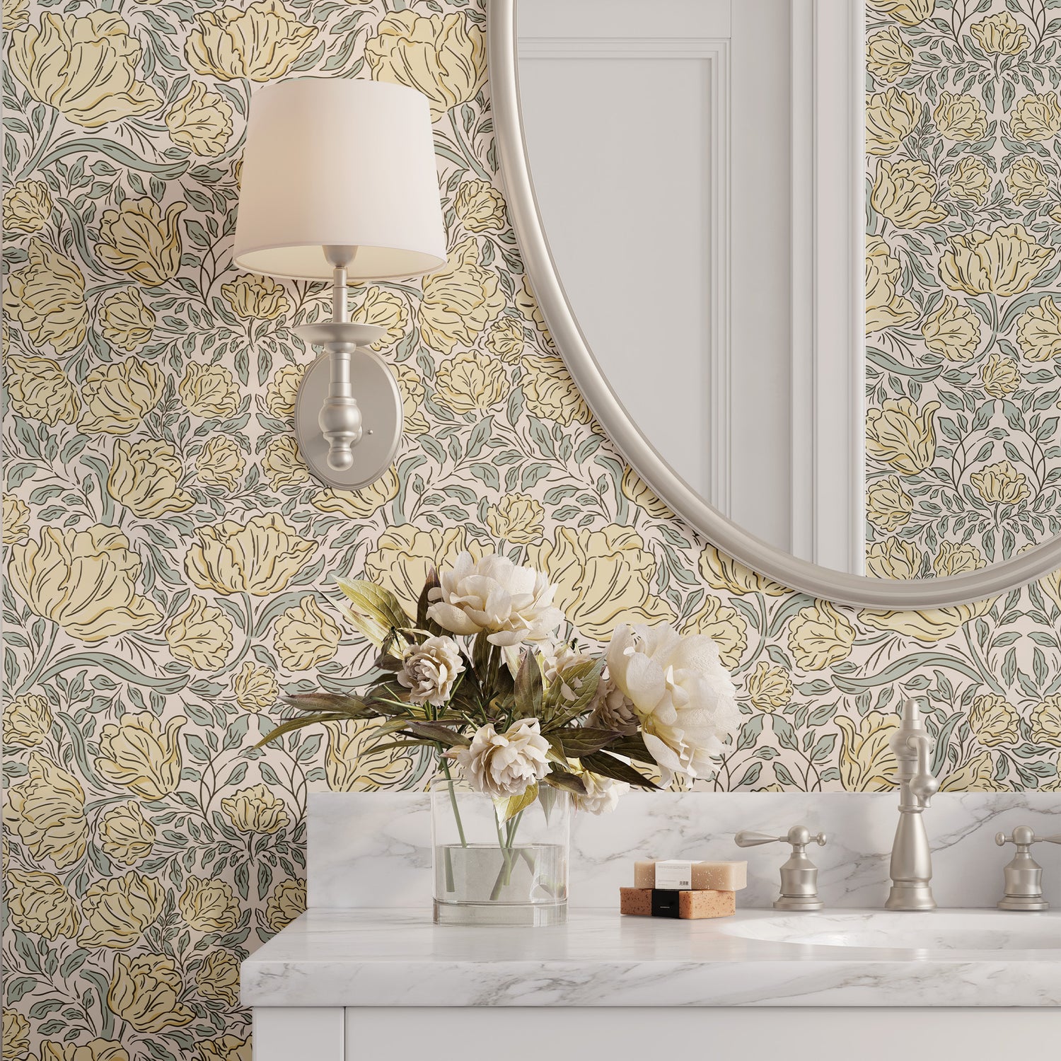 A styled bathroom scene featuring wallpaper with a vintage floral pattern in muted yellow, gray, and beige tones. A white marble vanity with a chrome faucet is visible at the bottom, along with a wall-mounted sconce, a large oval mirror, and a vase of white flowers.