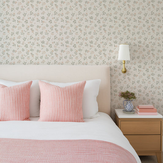 A styled bedroom scene featuring floral wallpaper with pink and green botanical motifs, a cream upholstered bed headboard, white bedding, pink striped pillows, a light wood nightstand, and a wall-mounted gold sconce.
